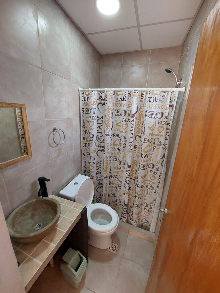un pequeño cuarto de baño con azulejos de color beige, un lavabo de piedra sobre una encimera de azulejos, un espejo circular, un inodoro, una papelera y una ducha con una cortina decorada con palabras como "amor", "paz" y "paz" en varios tipos de letra. en cabañas del sauce, sauce montrull, entre ríos