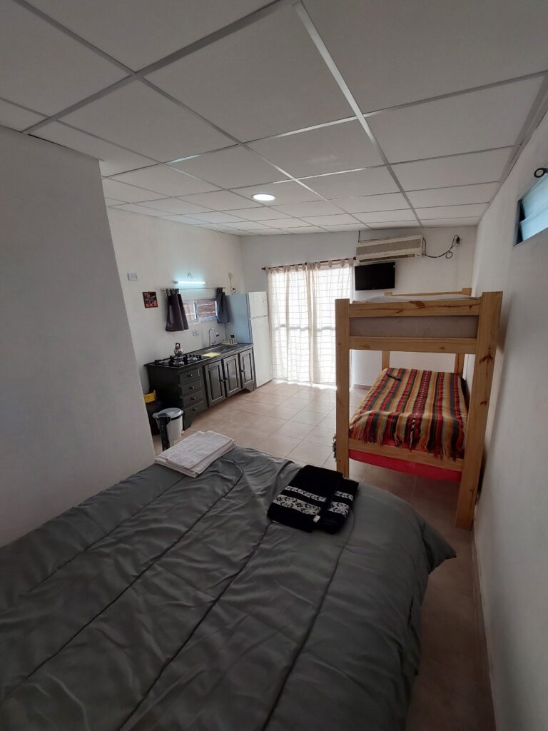 una habitación ordenada con una cama de matrimonio gris en primer plano y una litera de madera con ropa de cama de rayas de colores a la derecha. al fondo, hay una cómoda oscura, televisión, mini nevera, microondas y una ventana con cortinas transparentes que dejan entrar la luz del día. en cabañas del sauce, sauce montrull, entre ríos