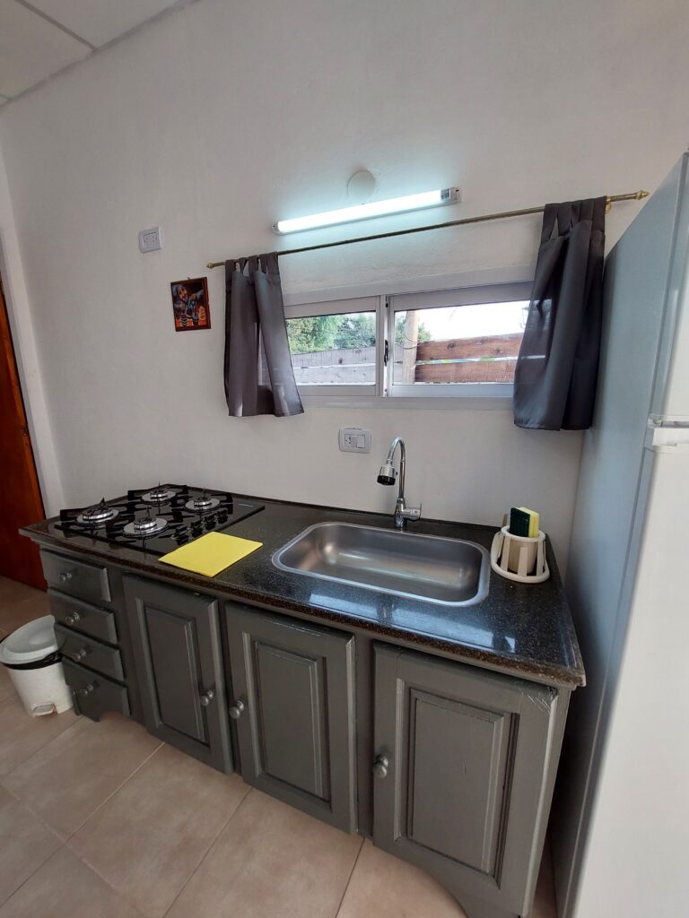 una cocina compacta presenta una encimera negra con un hornillo de gas, un fregadero, un dispensador de jabón y una tabla de cortar amarilla. arriba, dos pequeñas ventanas con cortinas grises y un fluorescente dejan pasar la luz natural. se ven armarios grises y un frigorífico. en cabañas del sauce, sauce montrull, entre ríos
