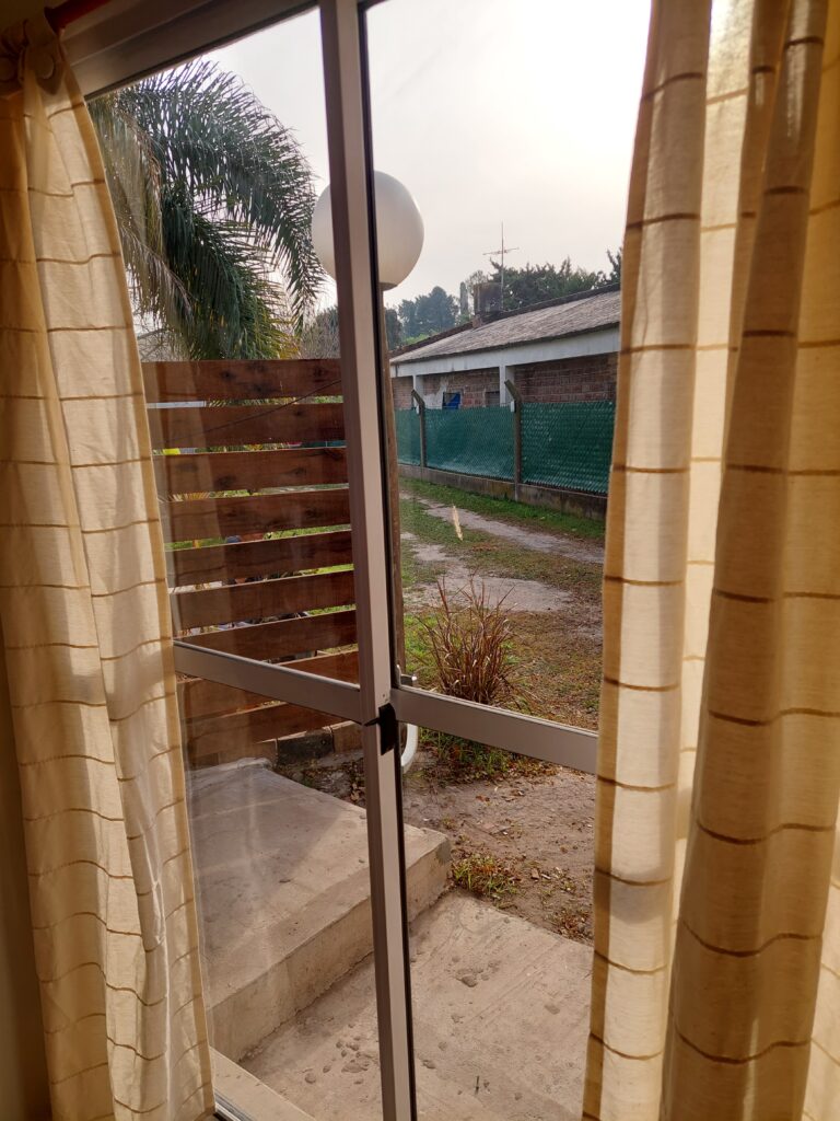 una puerta corredera de cristal con cortinas beige se abre a un pequeño escalón de hormigón y a un patio con un arbusto. en el exterior se ve una valla de madera, una malla verde y árboles bajo un cielo nublado. la luz del sol se filtra cálidamente a través de las cortinas. en cabañas del sauce, sauce montrull, entre ríos