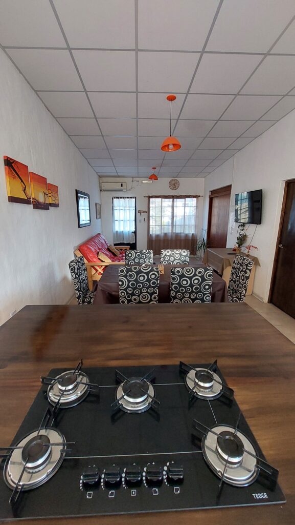 una cocina y un comedor modernos con un hornillo de gas de cuatro fuegos sobre una encimera de madera en primer plano. más allá hay una mesa de comedor con seis sillas estampadas, una sala de estar con sofás rojos, obras de arte en la pared, grandes ventanales y lámparas colgantes naranjas. en cabañas del sauce, sauce montrull, entre ríos