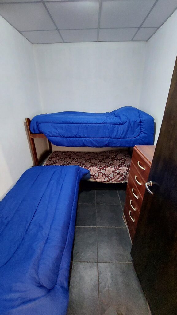 una habitación pequeña con suelo de baldosas negras, dos literas a lo largo de la pared izquierda la de arriba y la de abajo con edredones azules, y una cómoda de madera con tiradores plateados a la derecha. se ven paredes blancas y un techo de rejilla blanca. en cabañas del sauce, sauce montrull, entre ríos