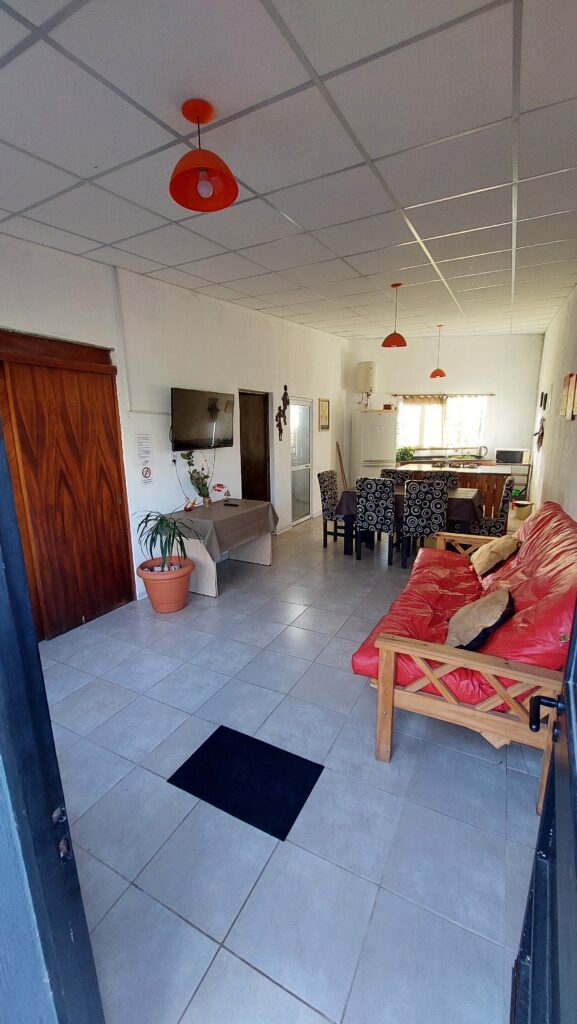 un luminoso salón con suelos de baldosas blancas, un sofá rojo con cojines, un televisor colgado en la pared, una maceta, una mesa de comedor de madera con sillas estampadas, puertas de madera y lámparas colgantes rojas en el techo. la luz del sol entra por una ventana al fondo. en cabañas del sauce, sauce montrull, entre ríos