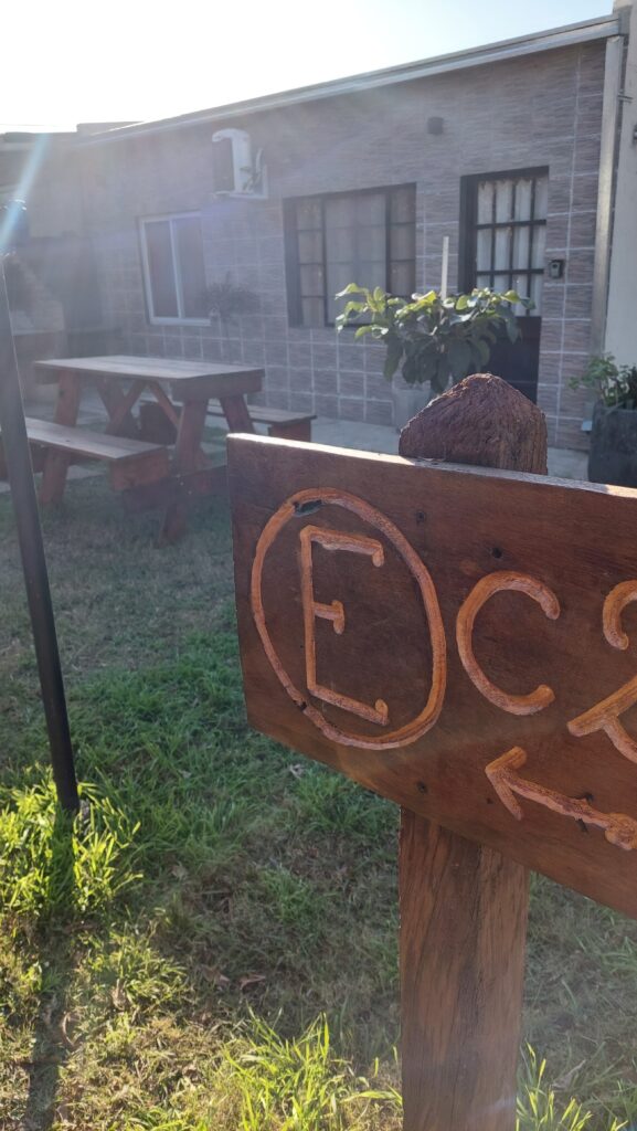primer plano de un cartel de madera con una letra e tallada y una flecha, delante de un patio cubierto de hierba. al fondo, una mesa de picnic de madera y un edificio de una sola planta con paredes de azulejos, ventanas y una puerta. la luz del sol proyecta largas sombras sobre la escena. en cabañas del sauce, sauce montrull, entre ríos