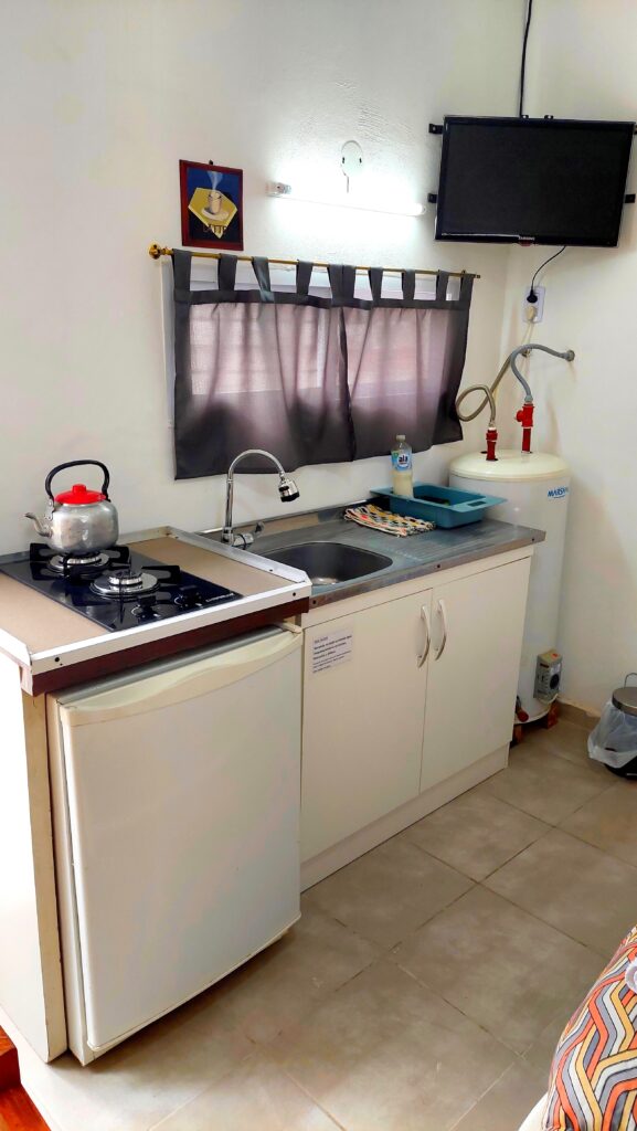 una cocina compacta con un pequeño frigorífico, cocina de gas de dos fuegos, hervidor de agua, fregadero, escurreplatos y jabón. encima del fregadero hay una ventana con una cortina gris. a la derecha hay un calentador de agua y un televisor de pared. el suelo es de baldosas beige. en cabañas del sauce, sauce montrull, entre ríos