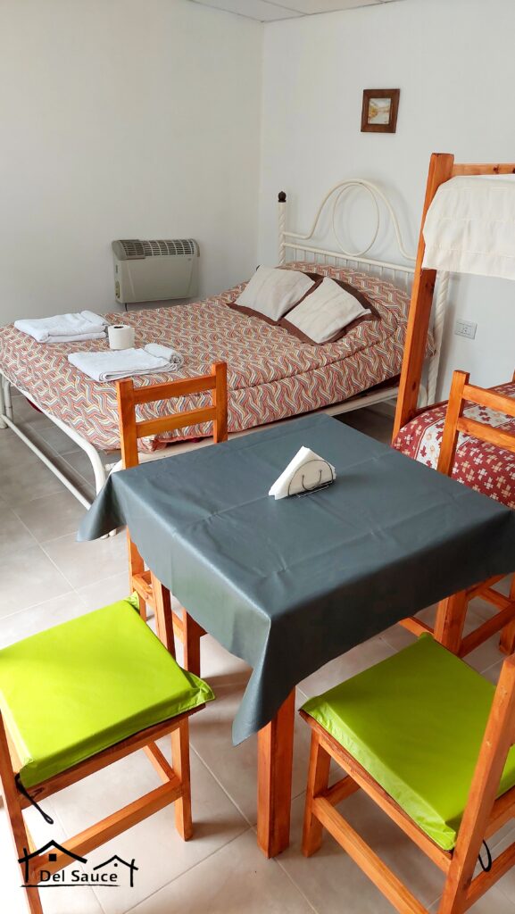 una acogedora habitación con una cama doble y una litera, ambas con ropa de cama estampada. en primer plano hay una mesita con un mantel oscuro y cuatro sillas de madera con cojines de color verde vivo. las toallas están dobladas sobre la cama; un cuadro enmarcado cuelga de una pared blanca. en cabañas del sauce, sauce montrull, entre ríos