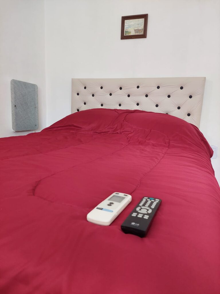 una cama pulcramente hecha con un edredón rojo y un cabecero crema empenachado. dos mandos a distancia, uno blanco y otro negro, descansan sobre la cama. hay un pequeño cuadro enmarcado en la pared blanca encima de la cama y un respiradero o panel en la pared izquierda. en cabañas del sauce, sauce montrull, entre ríos